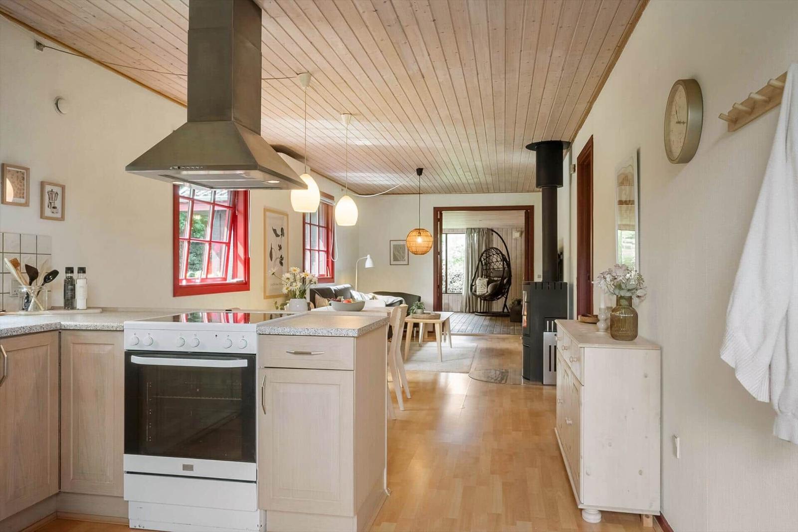 Kitchen with stove and oven, wooden ceiling, red windows, view into living area with seating.