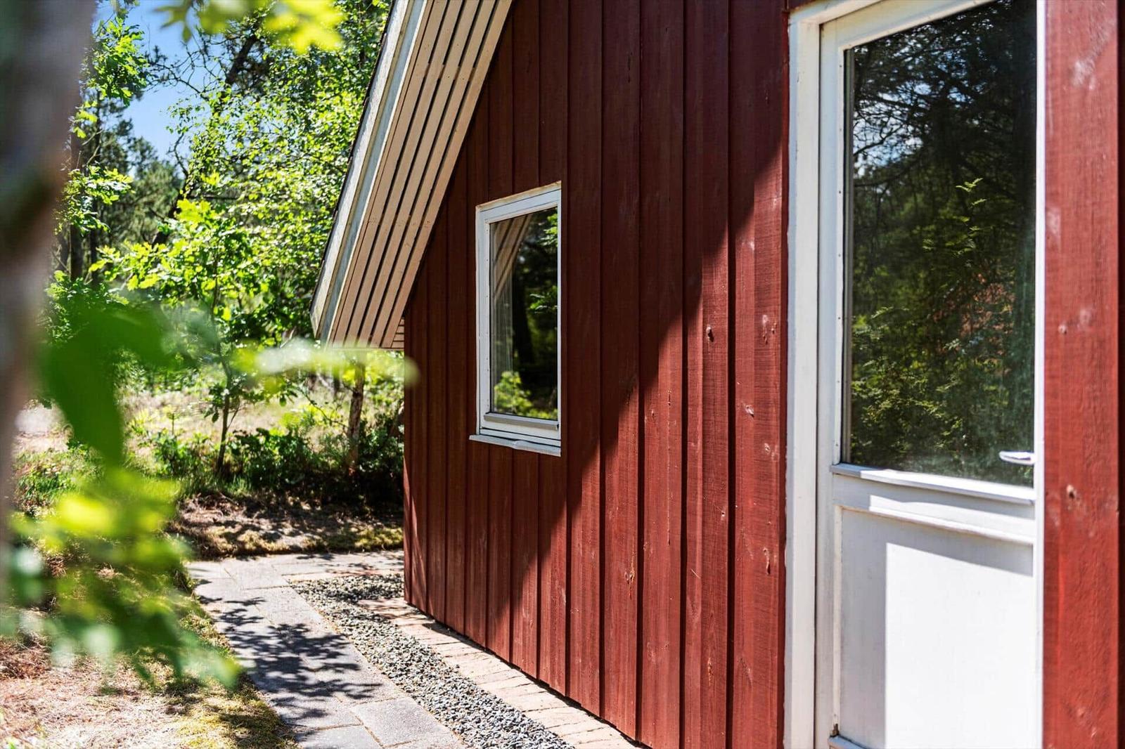 Rotes Holzhaus mit weißer Tür und Fenster, umgeben von Bäumen und einem Kiesweg.