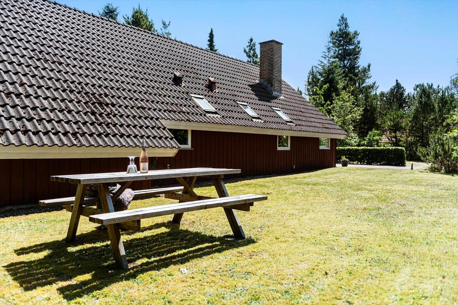 Holztisch mit Flasche auf Rasen vor rotem Holzhaus mit Schindeldach und Schornstein.