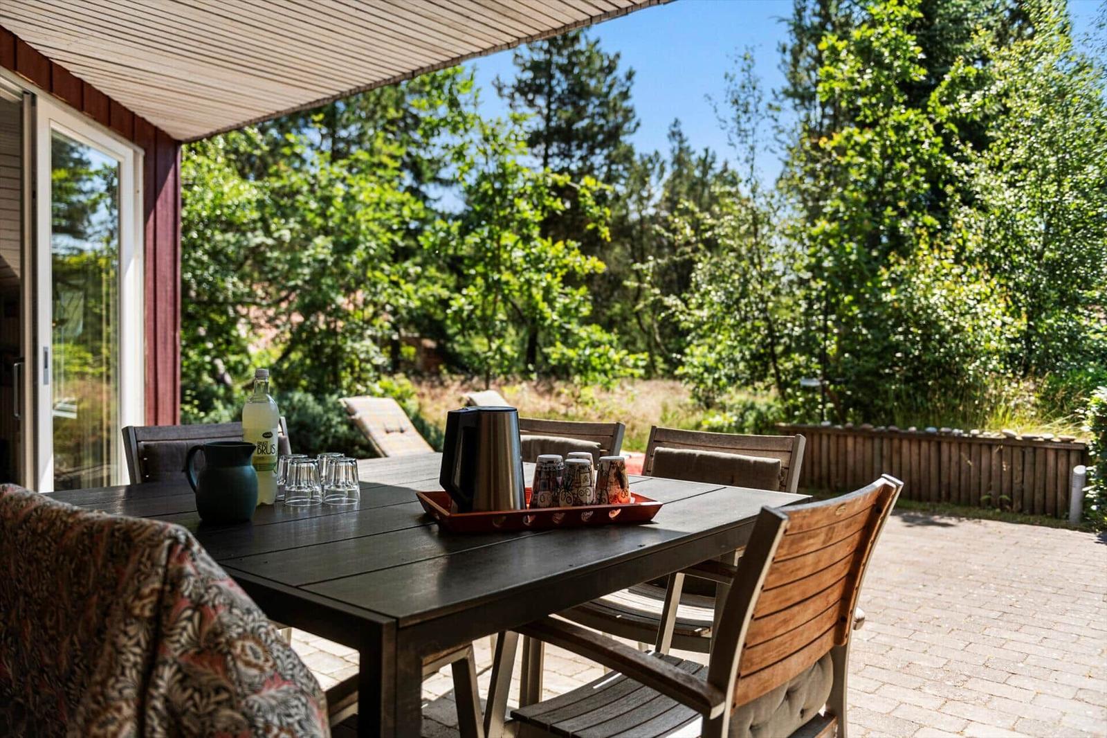 Terrasse mit Tisch, Stühlen und Grillbereich. Hintergrund: grüner Garten und Bäume.