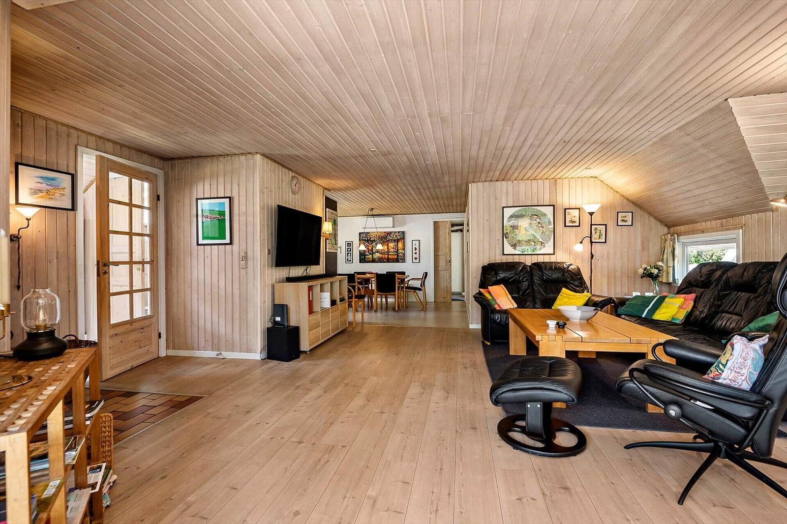 Wohnbereich mit Holzdecke, Sofa, Couchtisch und Fernseher. Im Hintergrund ist das Esszimmer.
