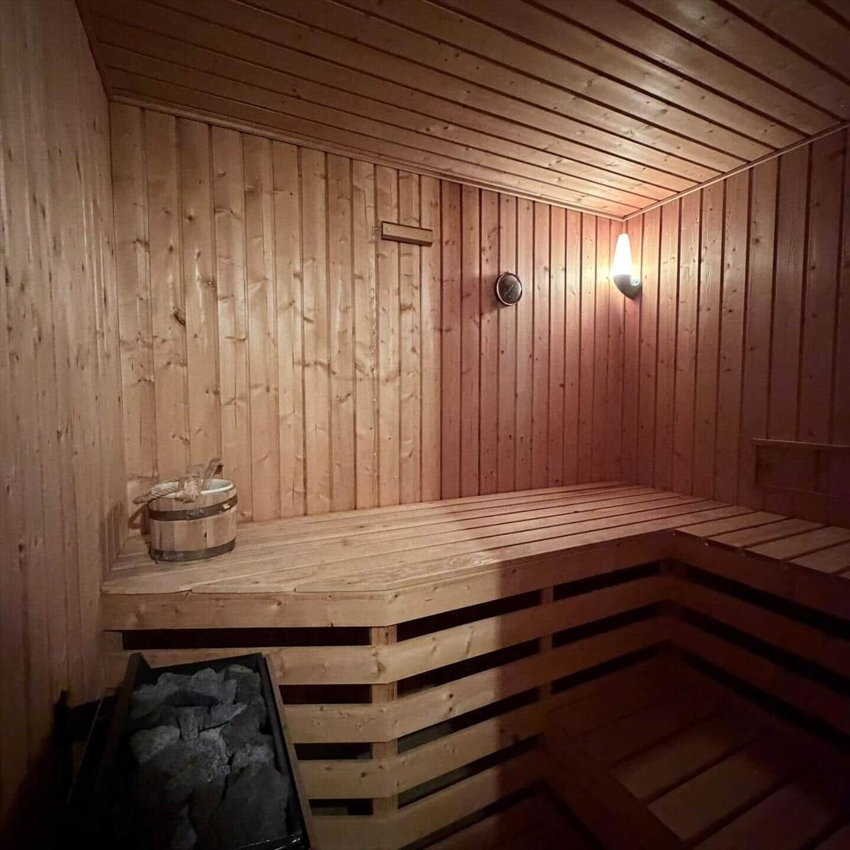 Holzsauna mit Sitzbank, Körbchen und Steinkasten. Wanduhr und Wandlampe sichtbar.