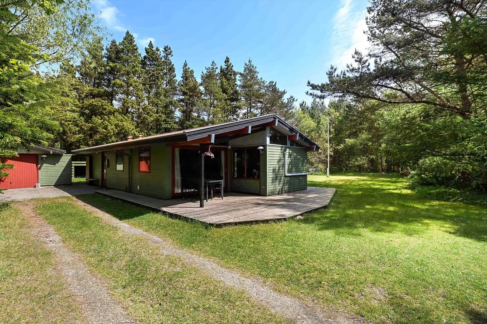 Grünes Ferienhaus mit Terrasse, umgeben von Wald und Grasfläche.