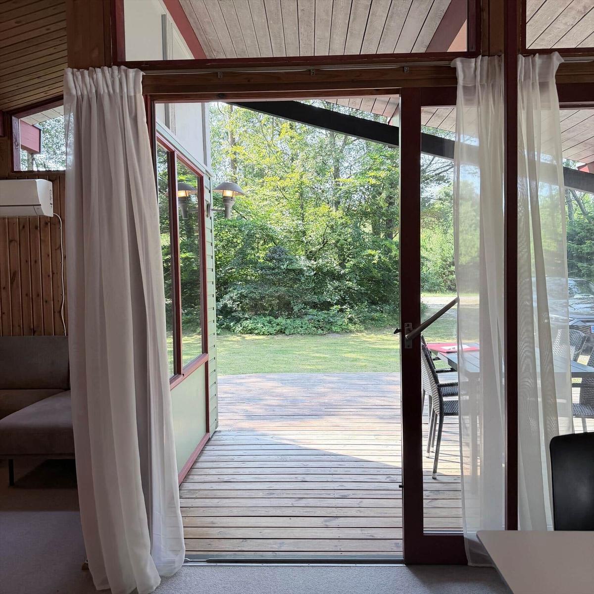 Durch die Glastüren führt ein Holzdeck in den Garten. Draußen steht ein Tisch mit Stühlen.
