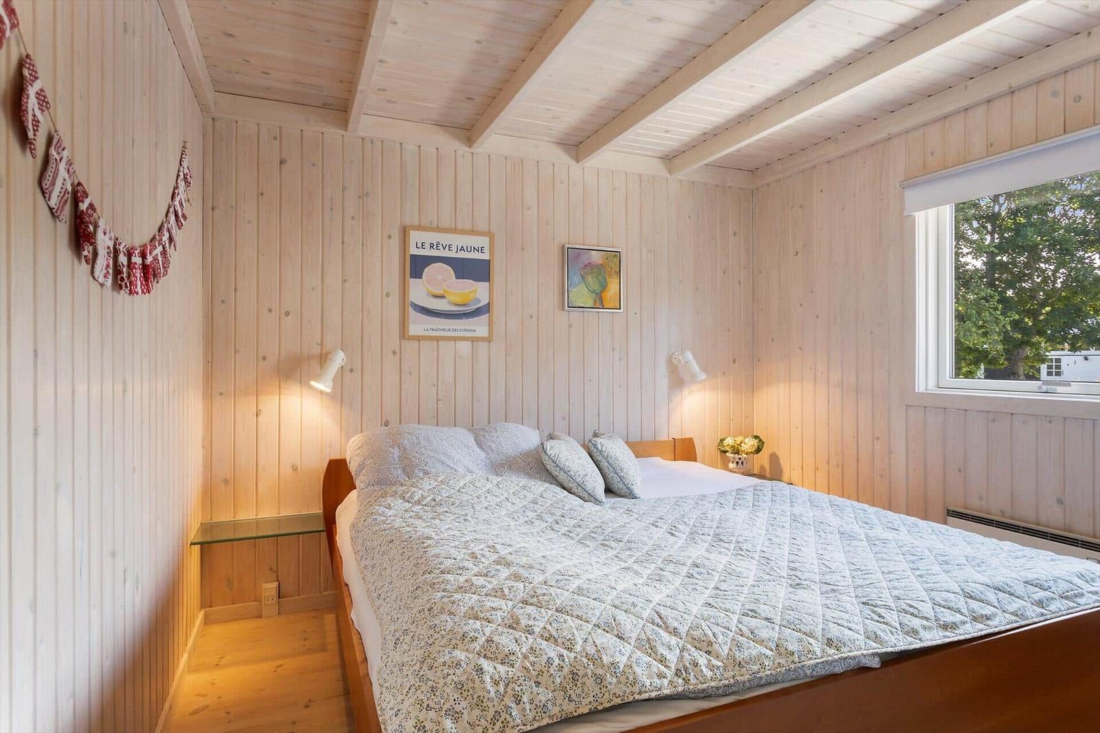 Schlafzimmer mit Holzwänden, Bett und Fenster. Wandbild und Girlande sichtbar.