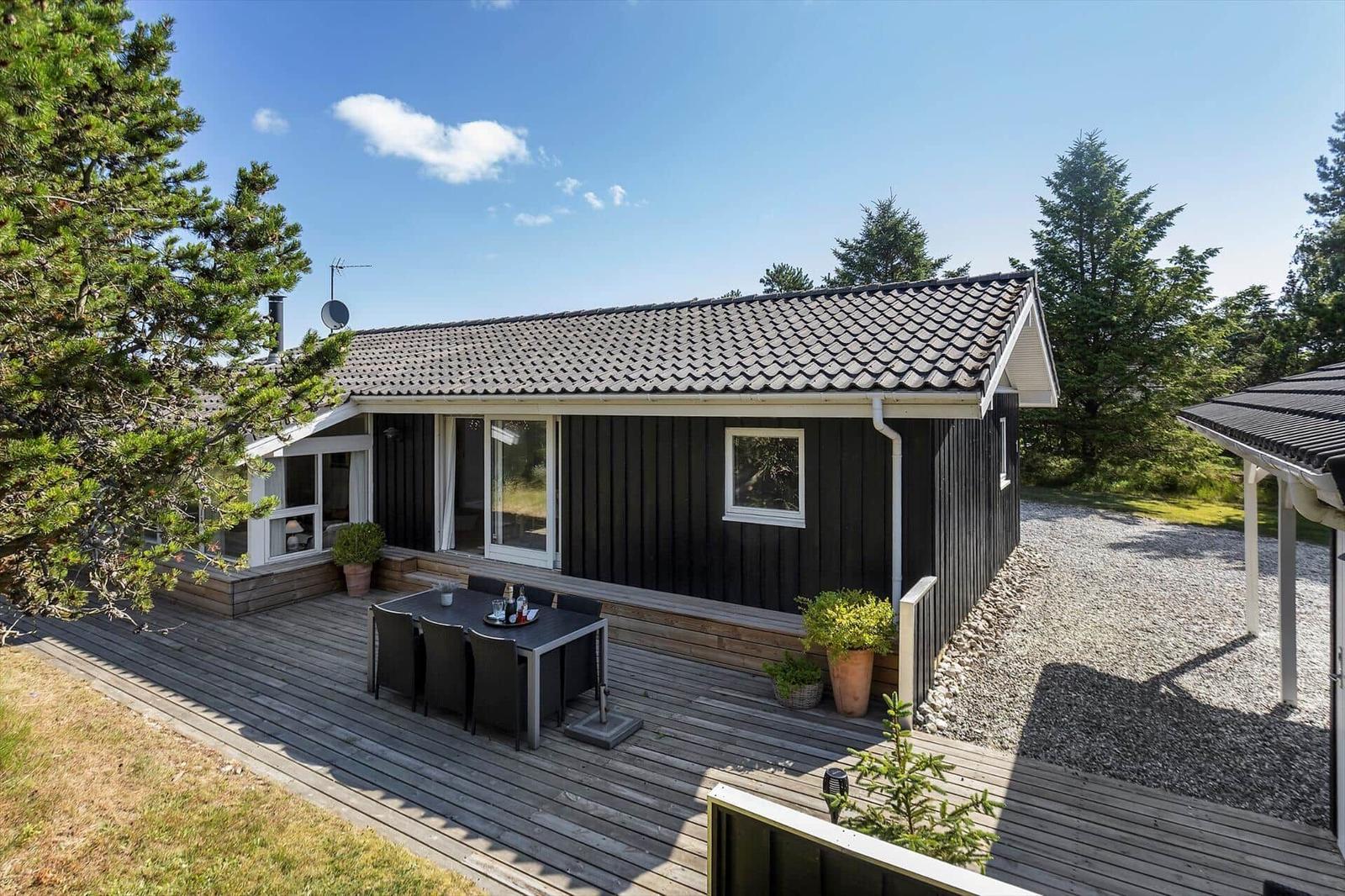 Ein Haus mit Holzterrasse, Gartentisch und Pflanzen unter blauem Himmel.