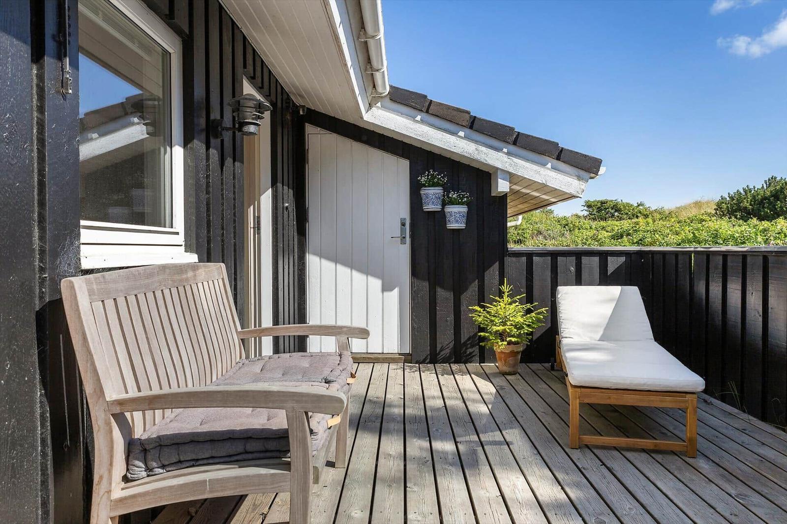 Terrasse mit Holzbank und Liegestuhl, schwarze Fassade, Pflanzen und klarer Himmel.