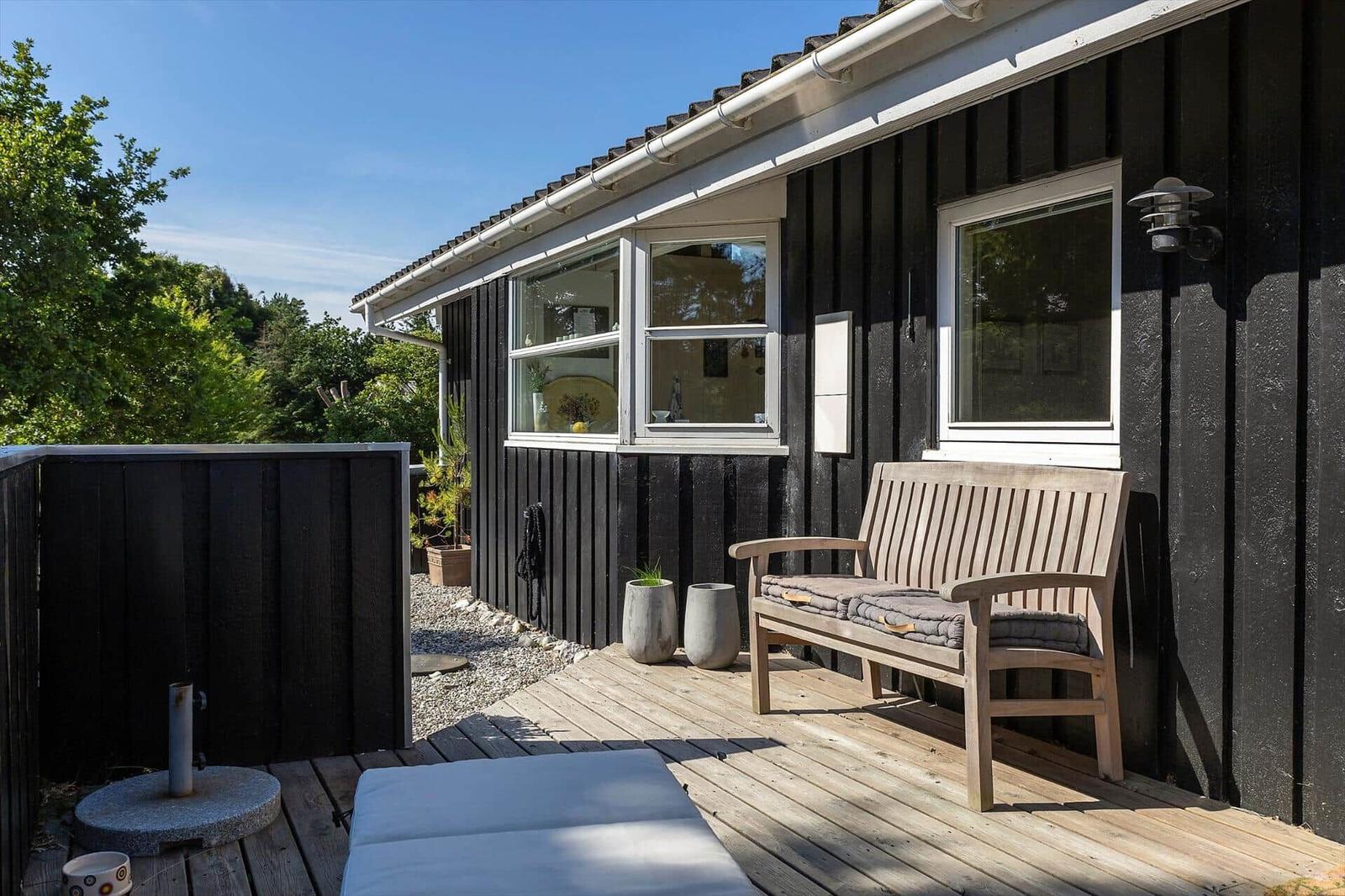 Terrasse mit Holzbank und Liege vor schwarzem Haus mit Fenstern.