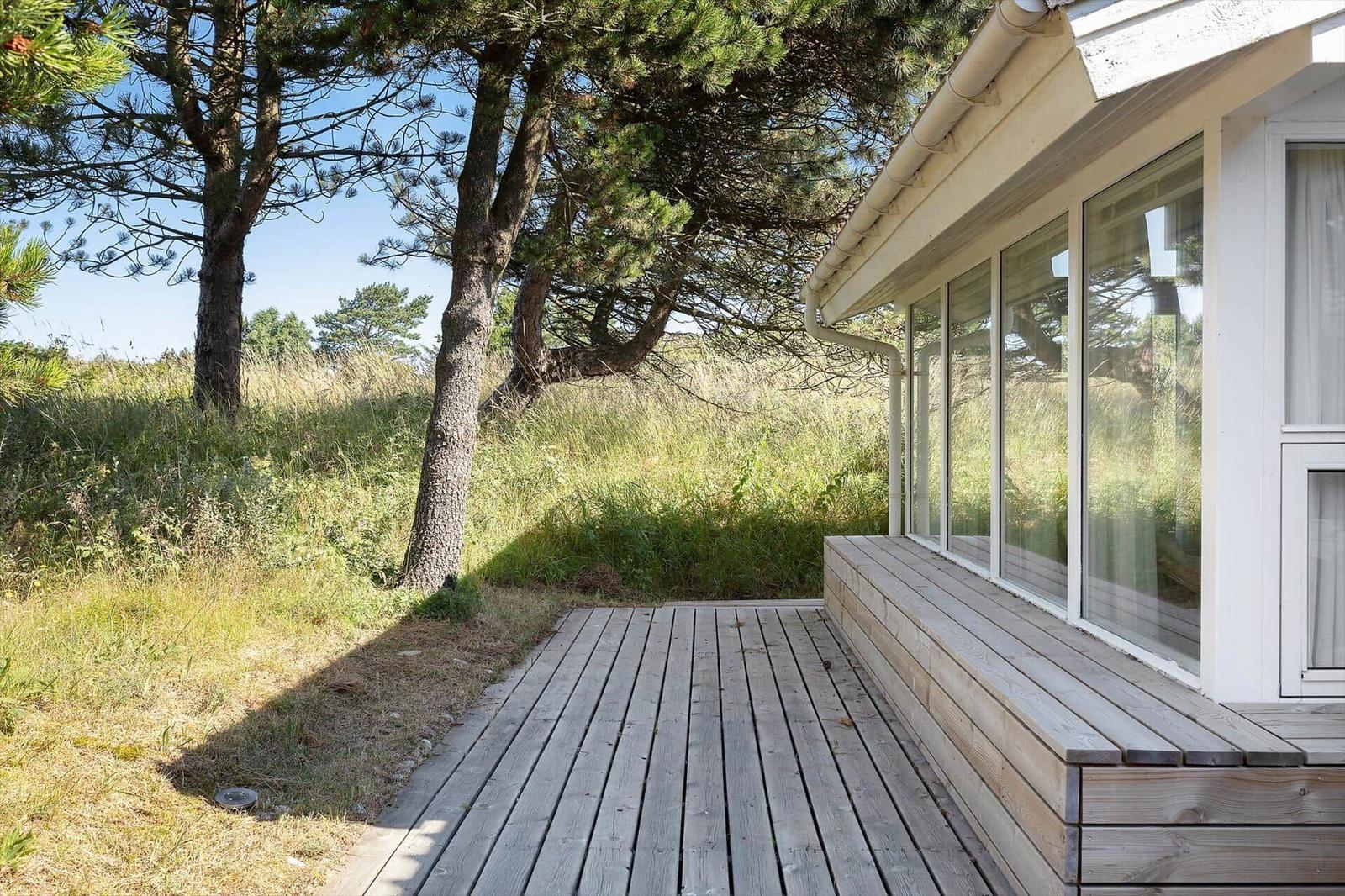Holzterrasse neben einem Haus mit großen Fenstern und Bäumen im Hintergrund.