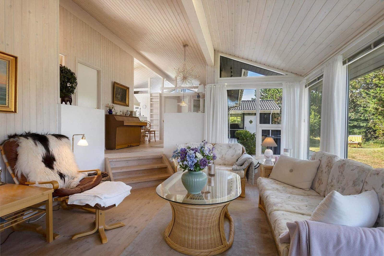 Wohnzimmer mit Sofa, Sessel und Glas-Tisch. Durchgang zur Küche. Große Fenster mit Blick auf Garten.