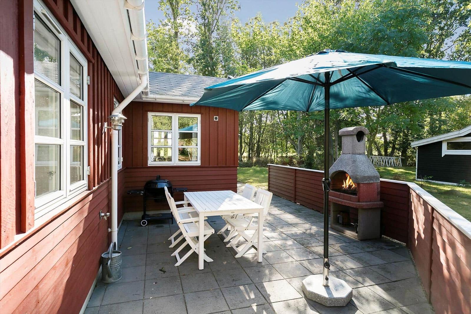 Terrace with table, chairs, umbrella, and grill. Background: forest and red wood walls.