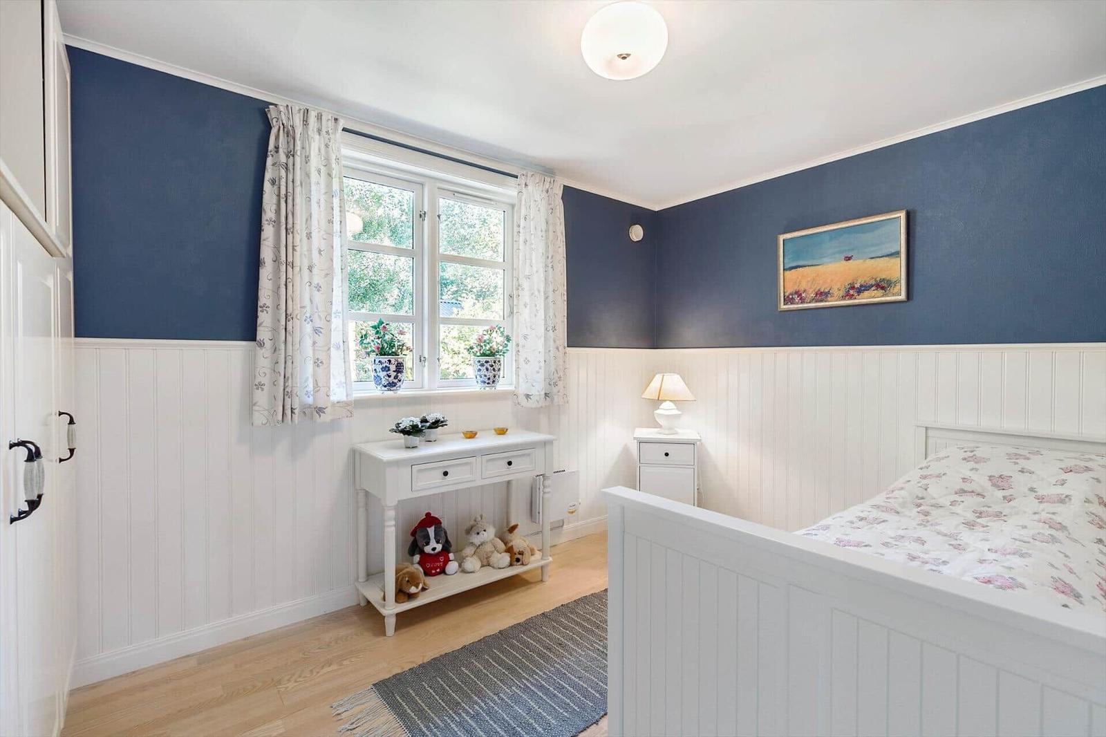 Bedroom with blue walls, white wainscoting, and wooden floor. Window with curtains, bed, and nightstand.