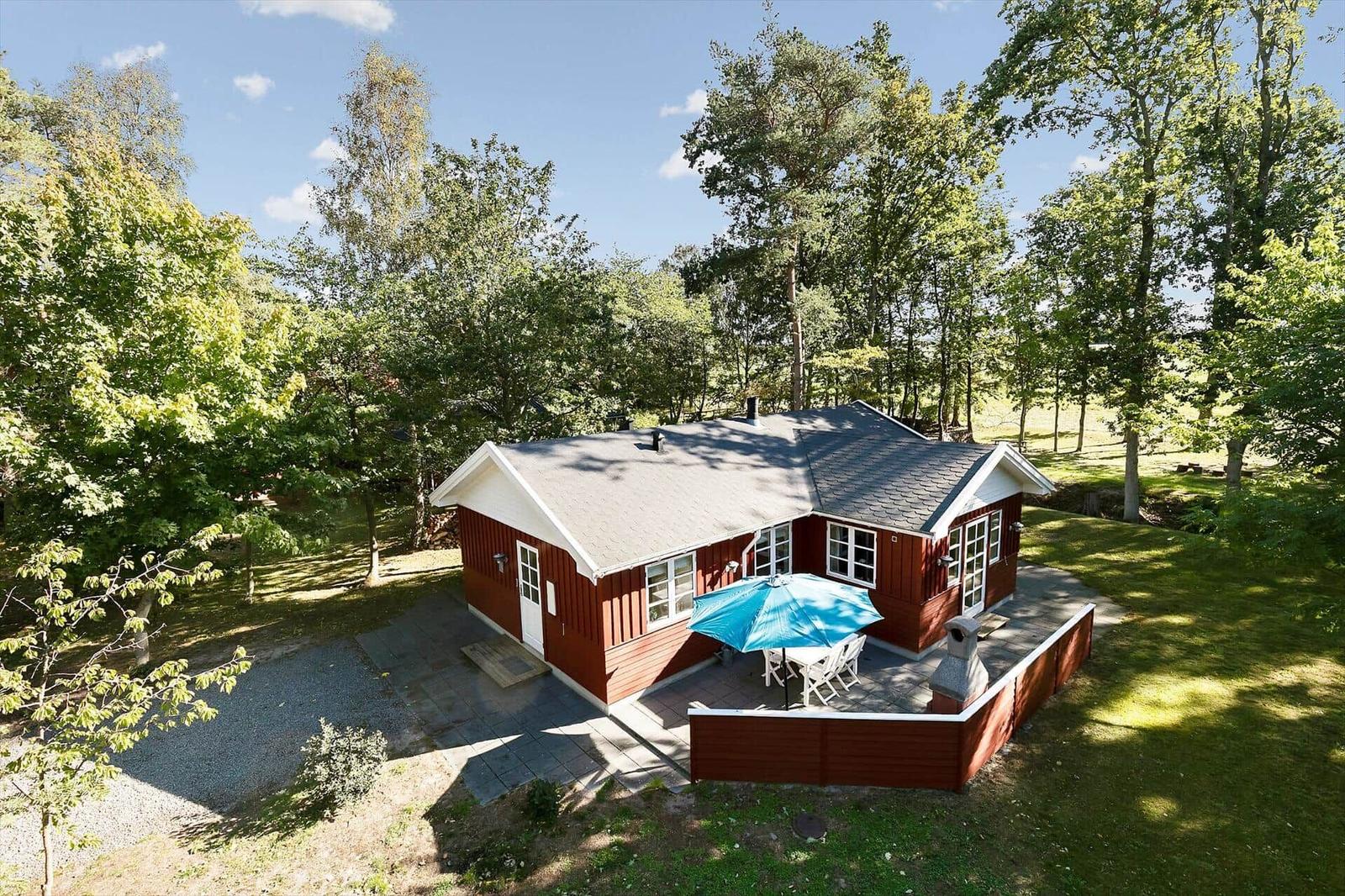 Red wooden house with terrace, blue umbrella, and garden surrounded by trees.