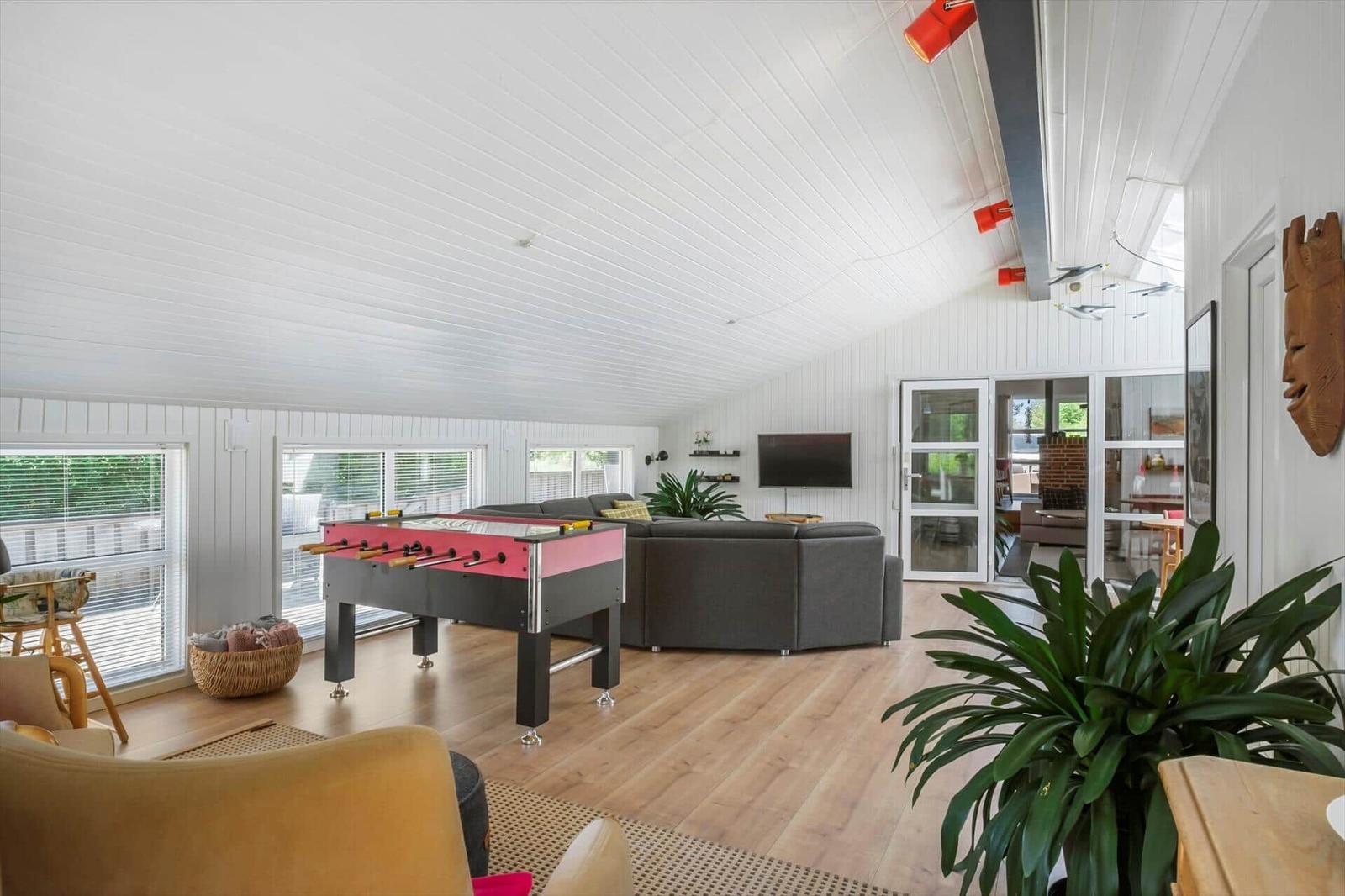 Wohnzimmer mit Tischfußball, Sofa und großen Fenstern. Holzboden und weiße Wände.