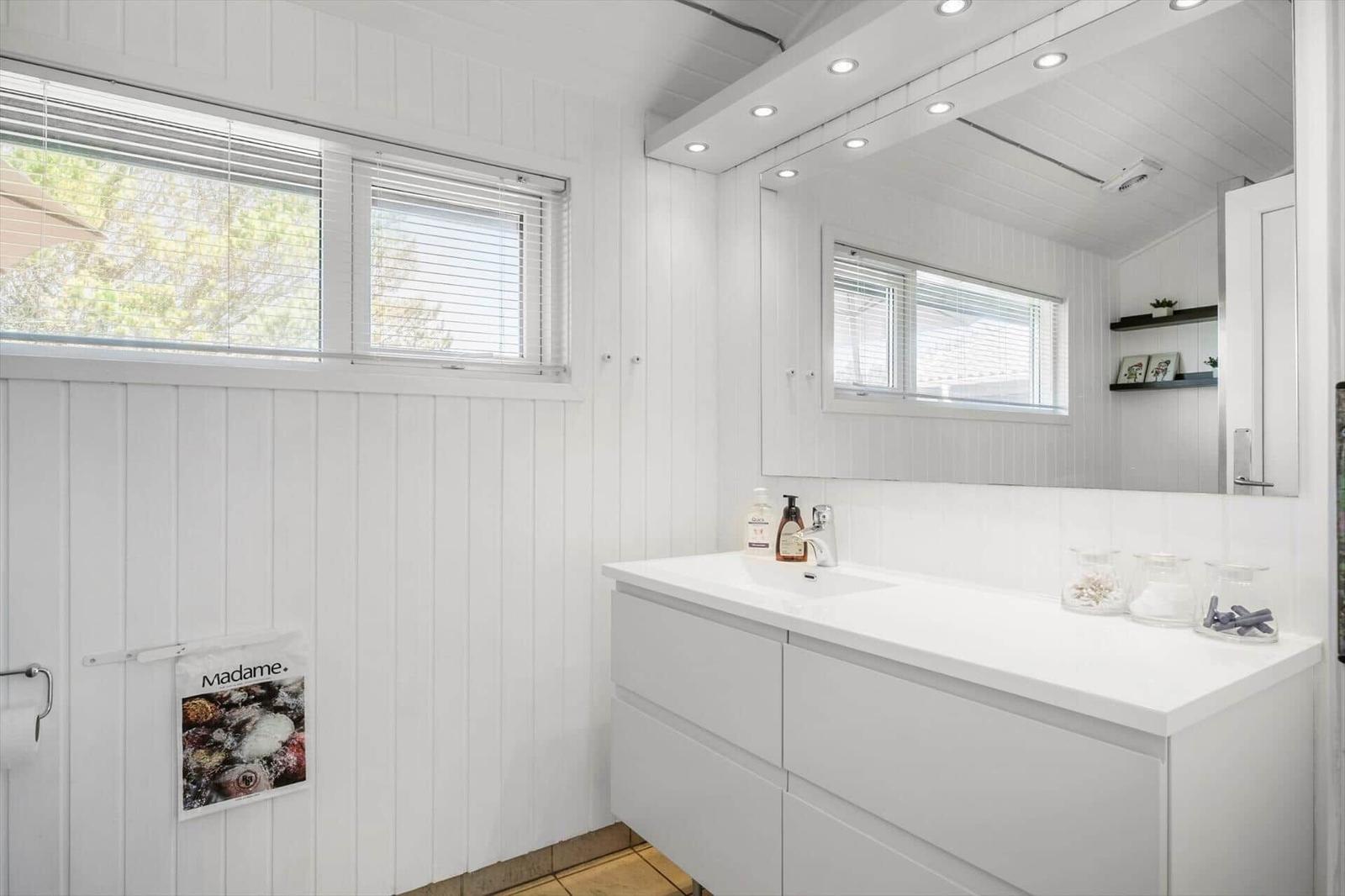 Badezimmer mit weißer Waschtischplatte, Spiegel und Fenster mit Jalousien.