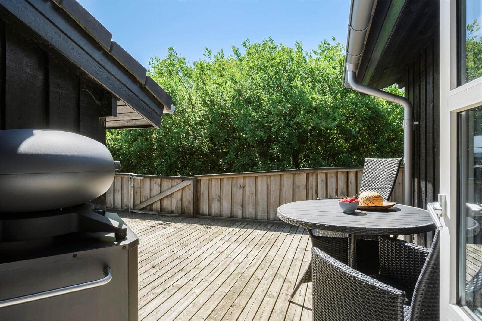 Terrasse mit Grill, Tisch und Stühlen. Holzgeländer und grüne Bäume im Hintergrund.