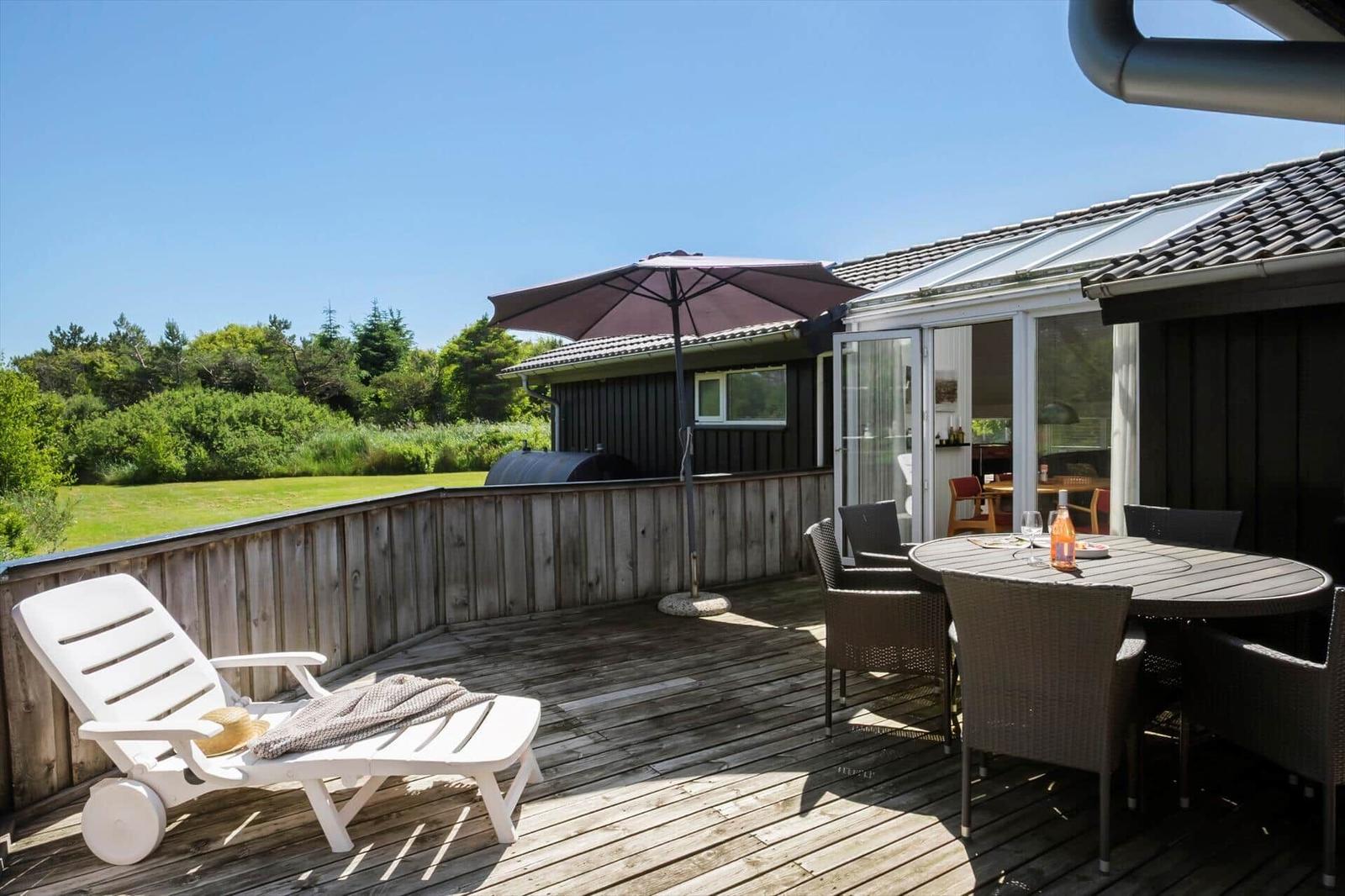 Terrasse mit Tisch, Stühlen, Sonnenschirm und Liegestuhl. Blick auf Grünfläche und Wald.