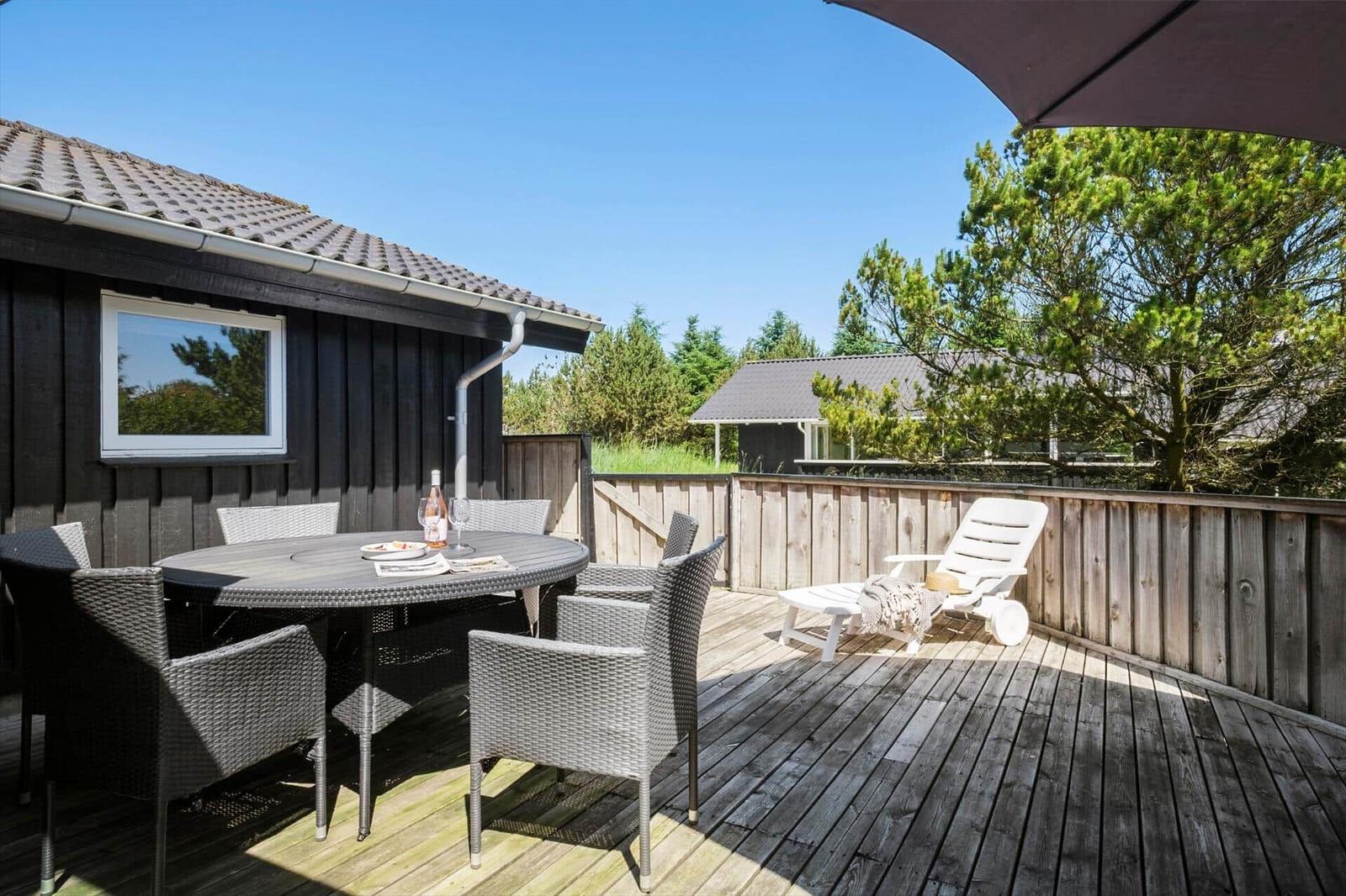 Terrasse mit Tisch, Stühlen und Liege. Holzdecke, Zaun und Bäume im Hintergrund.