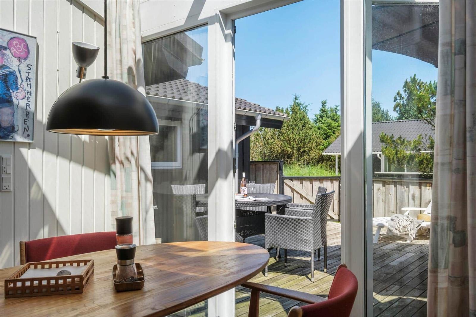 Innenraum mit Holztisch und roten Stühlen. Durch Glasfenster ist eine Terrasse mit Gartenmöbeln sichtbar.