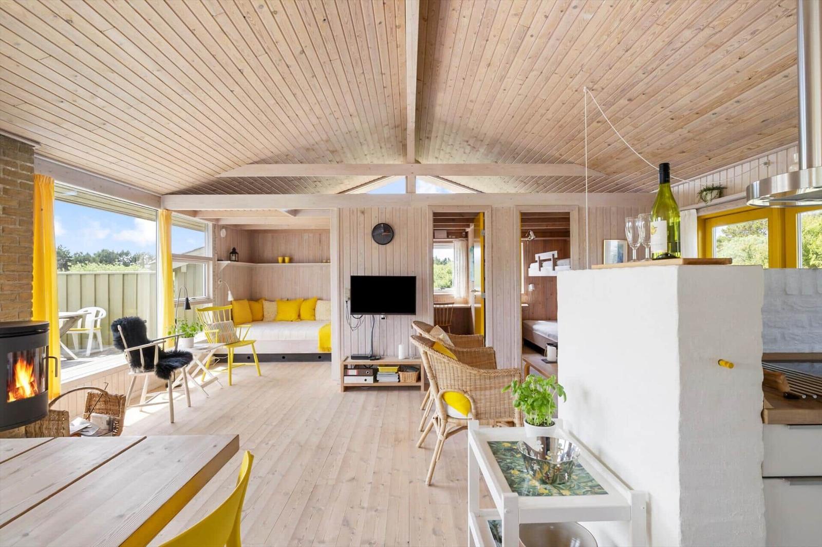 Wohnraum mit Holzdecke, gelben Sitzgelegenheiten und Kamin. Küche mit Arbeitsplatte und Tisch.