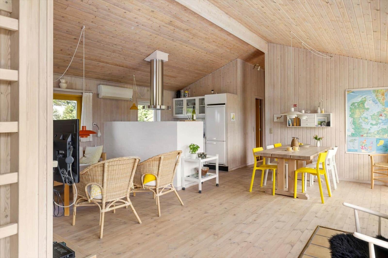 Wohnküche mit Holzdecke, Esstisch und gelben Stühlen. Kühlschrank und Dunstabzug sichtbar.