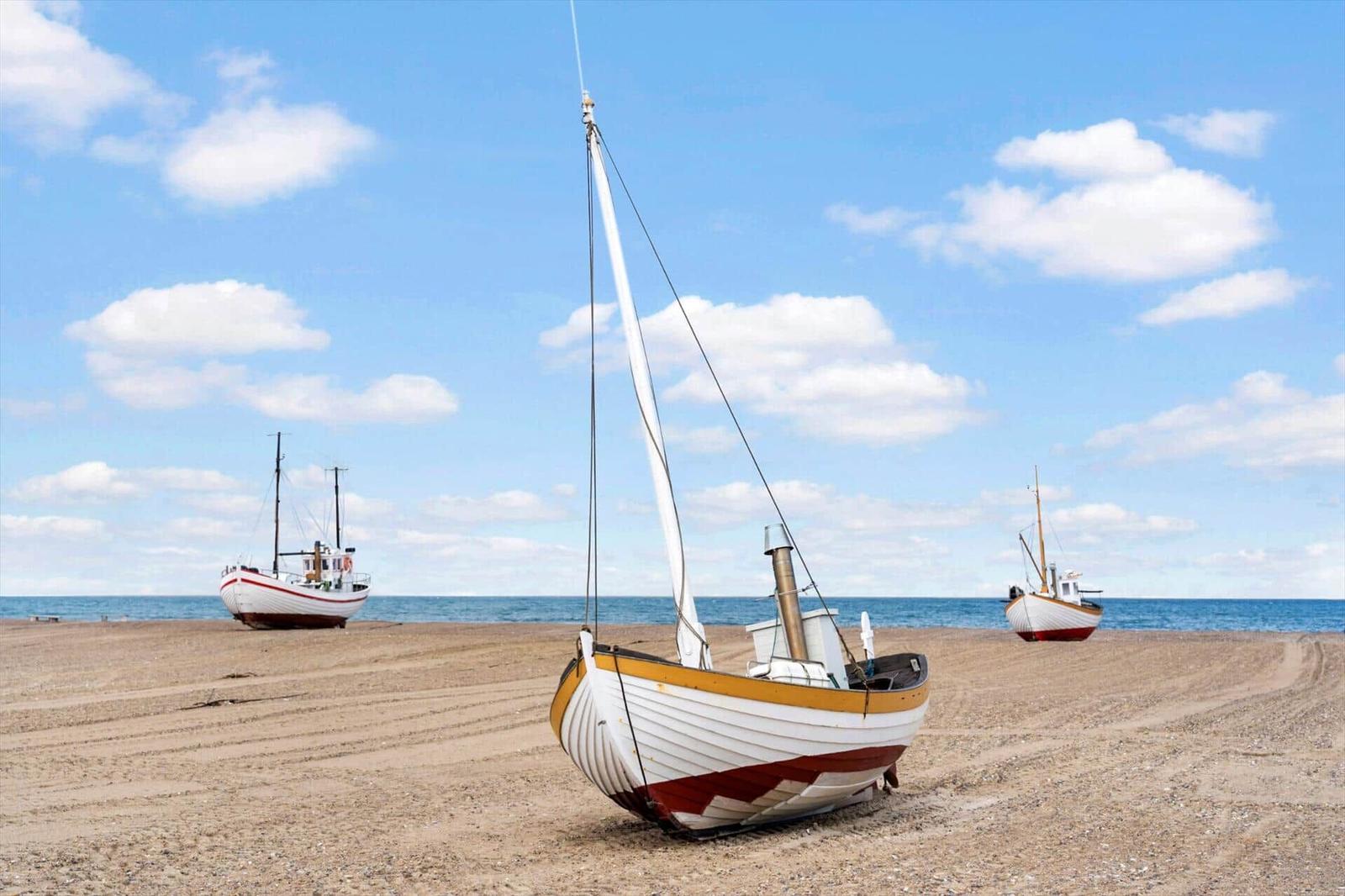 Drei Boote stehen auf einem sandigen Strand vor dem Meer unter blauem Himmel mit weißen Wolken.