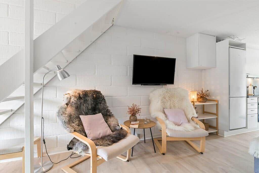 Two chairs with fur covers and cushions are placed in front of a wall with a TV and kitchen area.