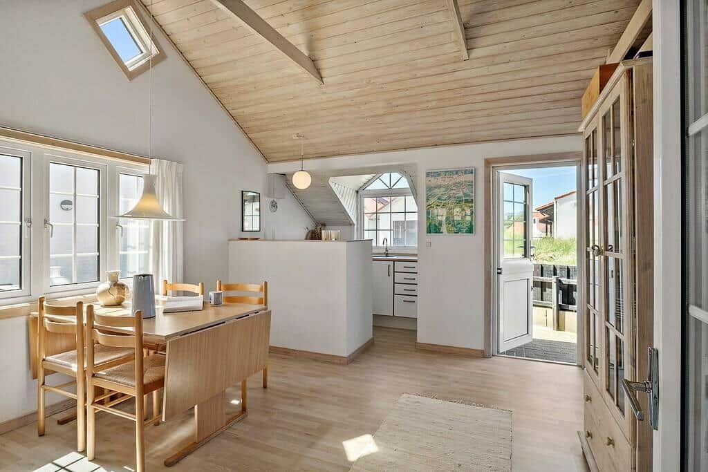 Dining room with wooden table, chairs, and windows. Kitchen and balcony are visible.