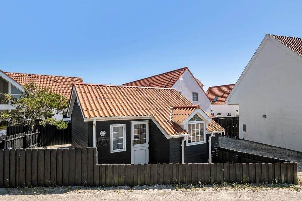 A small house with dark siding and red roof, surrounded by a wooden fence.