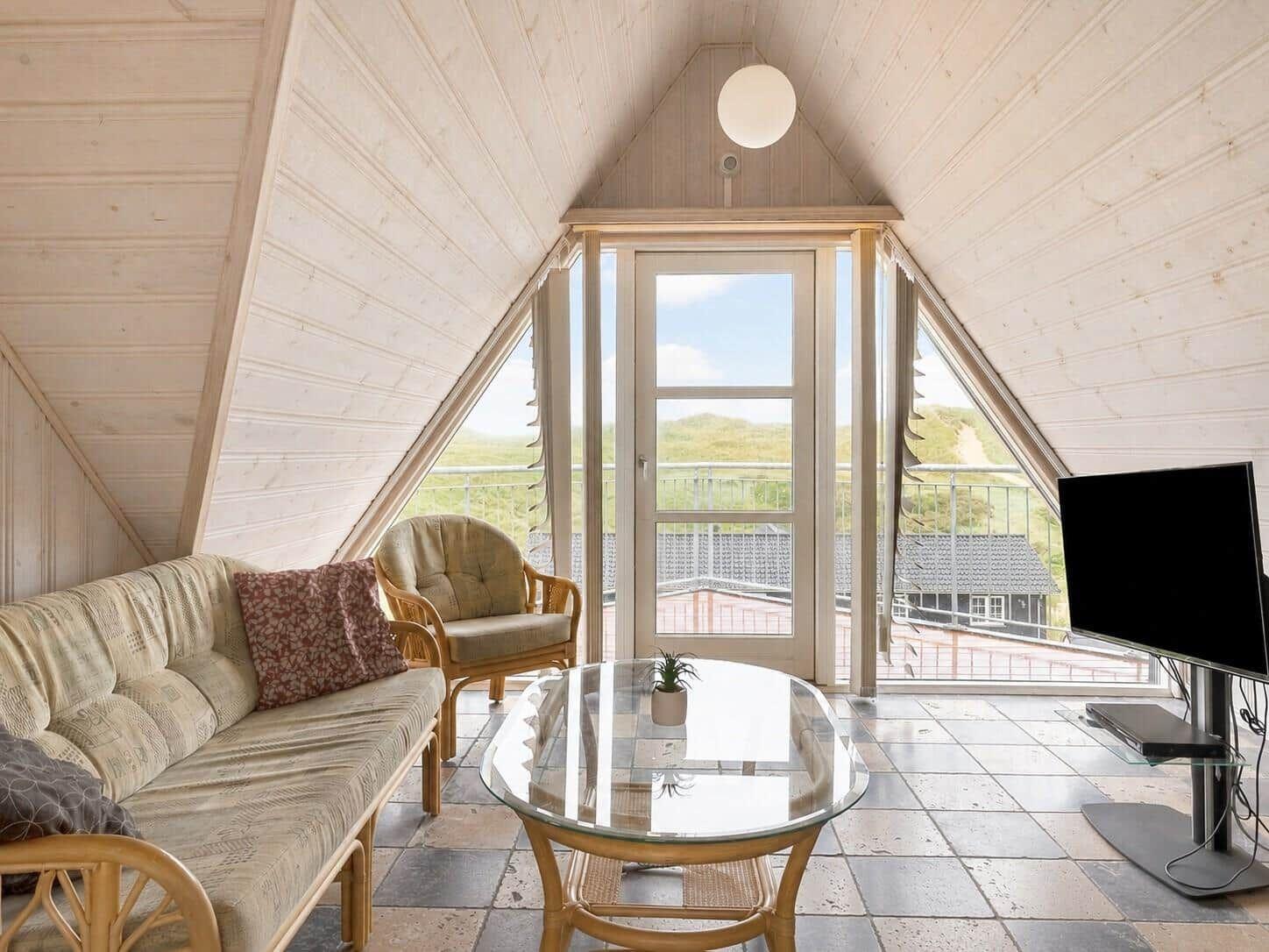 Wohnzimmer mit Sofa, Sessel, Glas-Tisch und TV. Durchgang zur Terrasse mit Blick auf Landschaft.