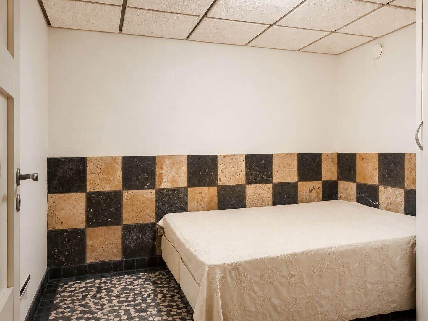 Schlafzimmer mit Bett, schwarz-weißem Mosaikboden und gemustertem Wandpaneel.