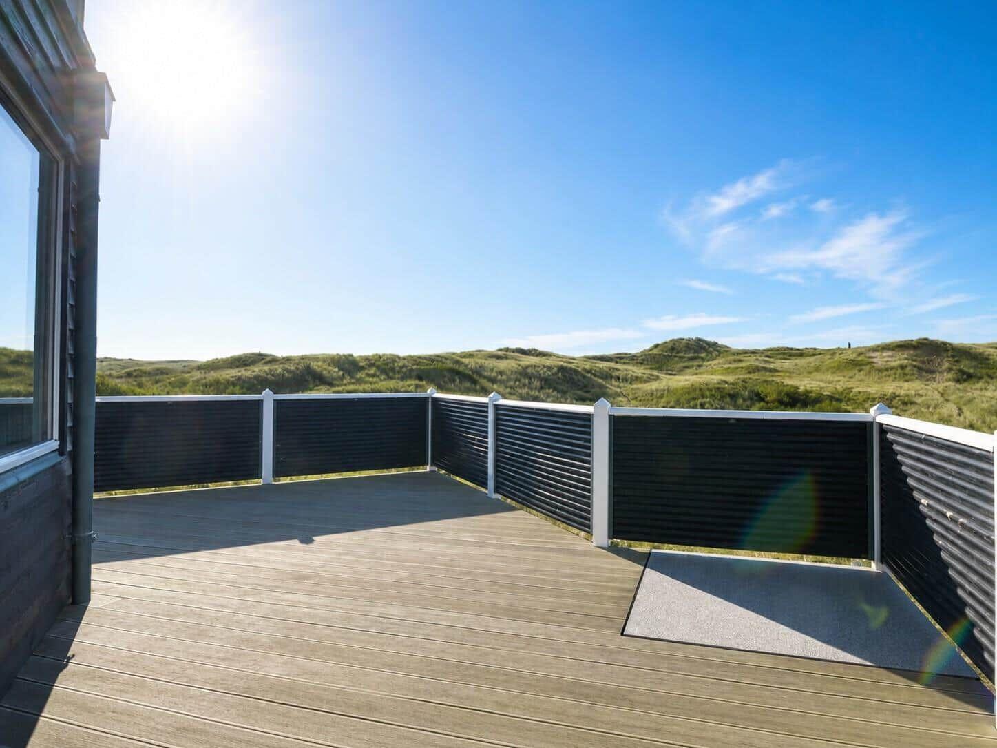 Terrasse mit Holzboden und schwarzer Brüstung, Blick auf grüne Dünen unter blauem Himmel.