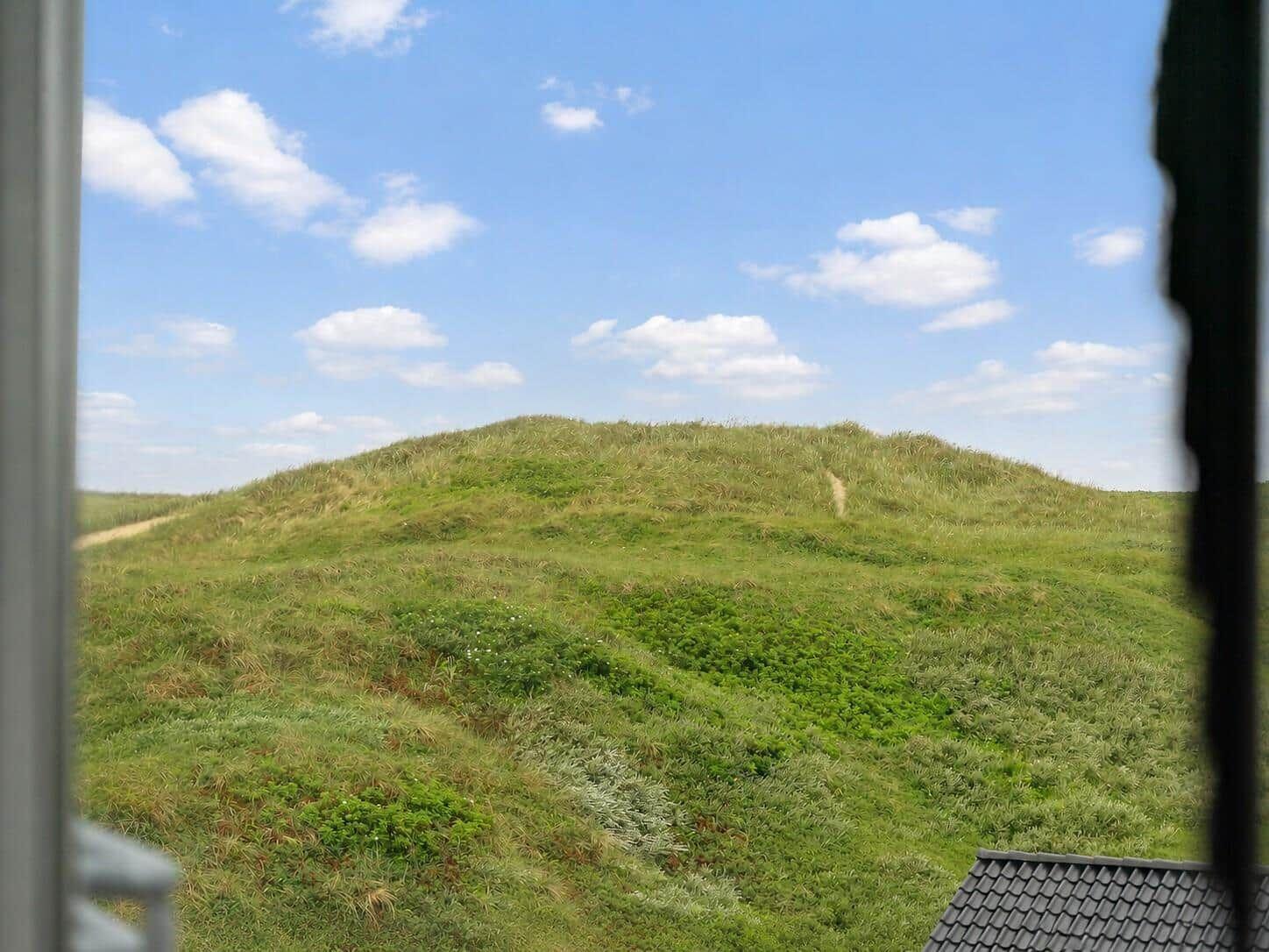 Grüne Dünen unter blauem Himmel mit weißen Wolken. Blick aus Fenster mit Teil des Daches.