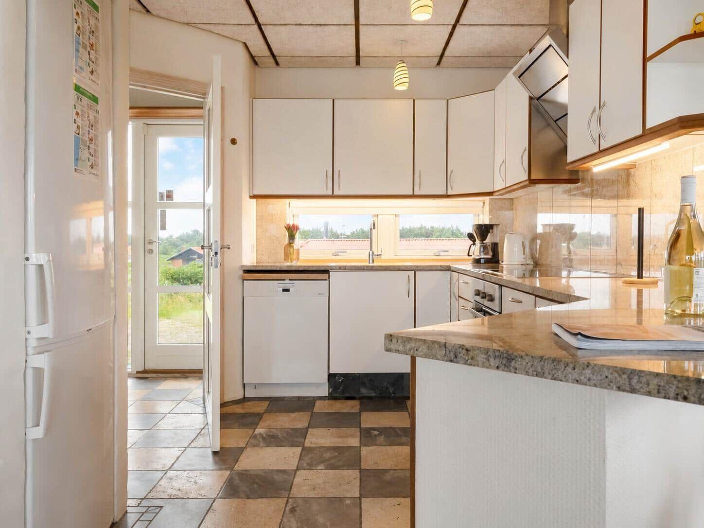 Küche mit weißen Schränken, Spüle und Fenster. Fliesenboden und Kühlschrank sichtbar.