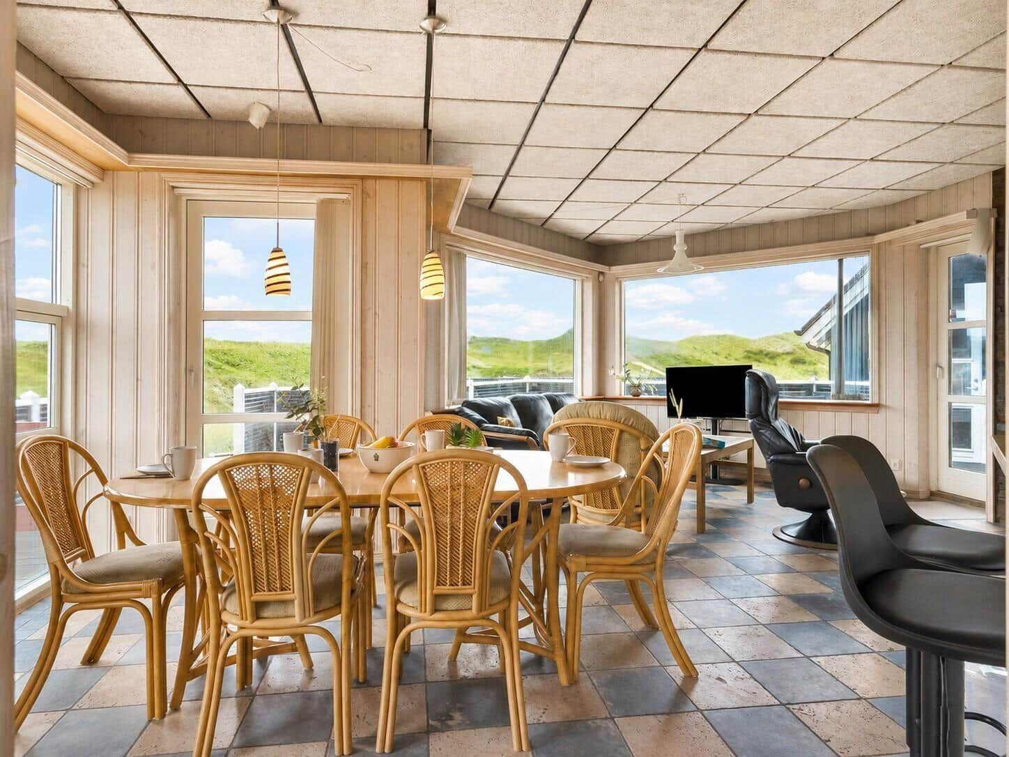 Essbereich mit Tisch und Stühlen, Sitzgelegenheiten und Fenstern mit Blick auf Grünflächen.