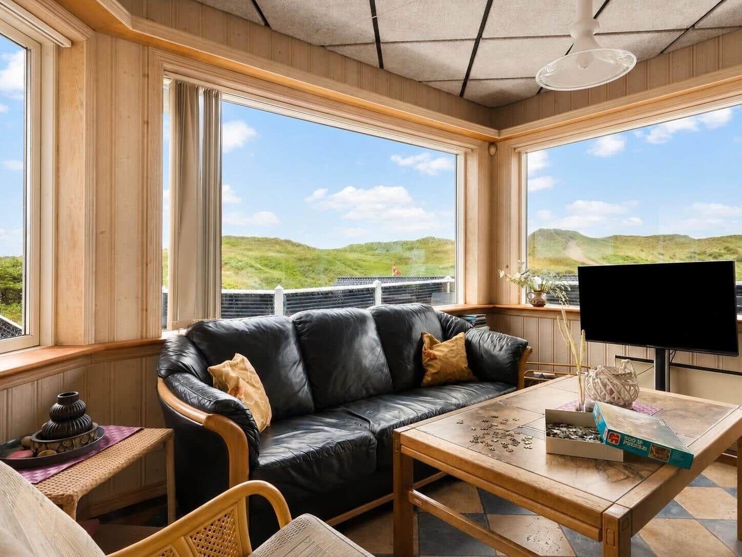 Wohnzimmer mit Sofa, Couchtisch und großen Fenstern mit Blick auf grüne Hügel.