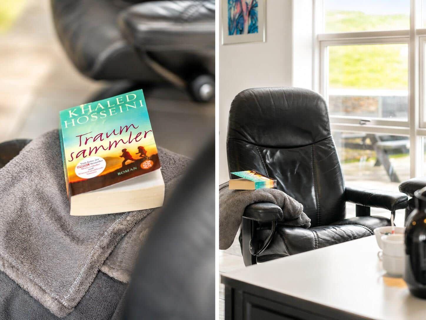 Ein schwarzer Sessel mit einem Buch und einer Decke. Durch das Fenster ist Grün zu sehen.