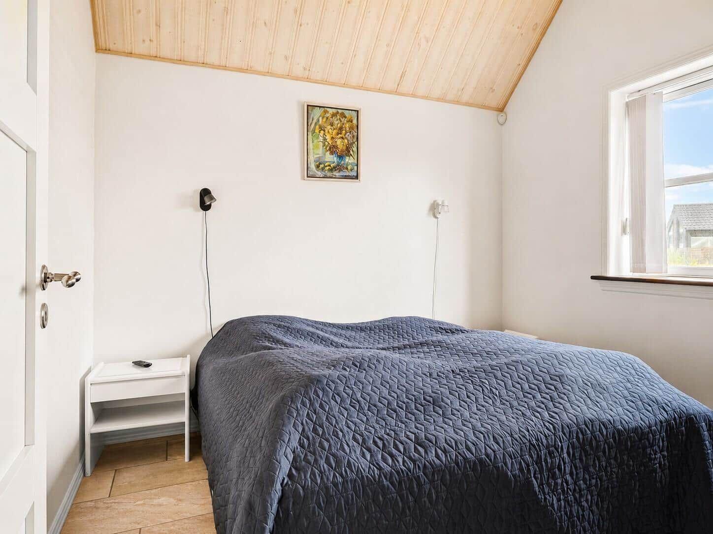 Schlafzimmer mit Bett, Nachttisch und Fenster. Holzdecke und Wandbild.