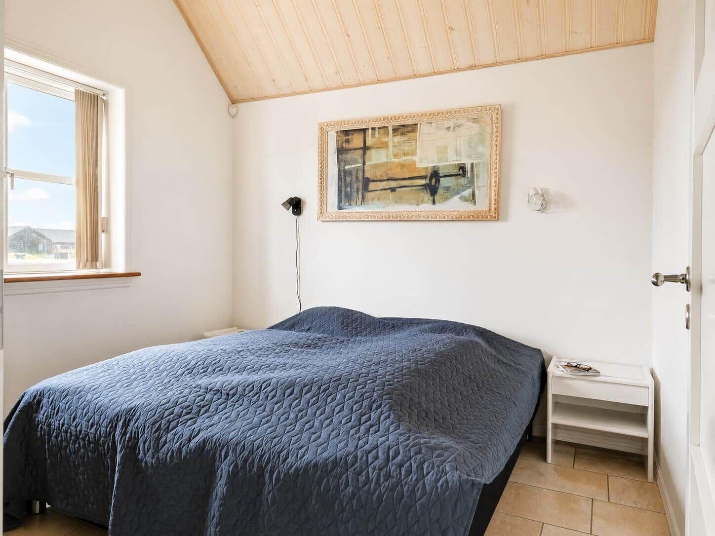 Schlafzimmer mit Bett, Fenster und Wandbild. Holzdecke und weißer Schrank.