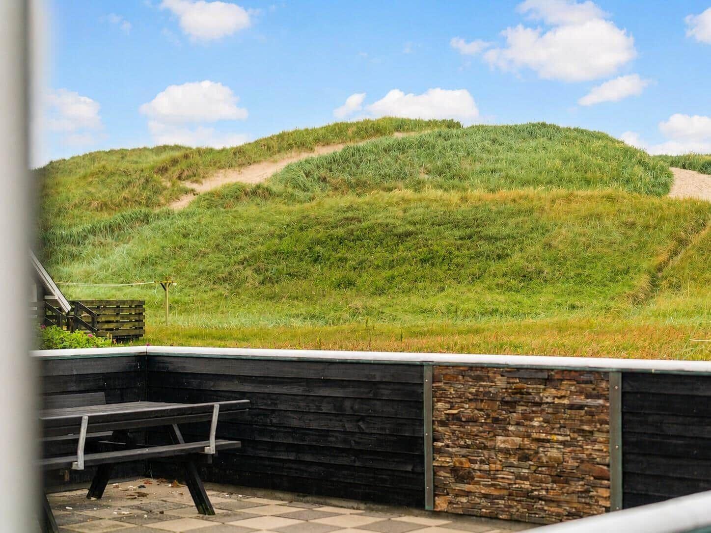 Terrasse mit Tisch und Blick auf grüne Dünen unter blauem Himmel.