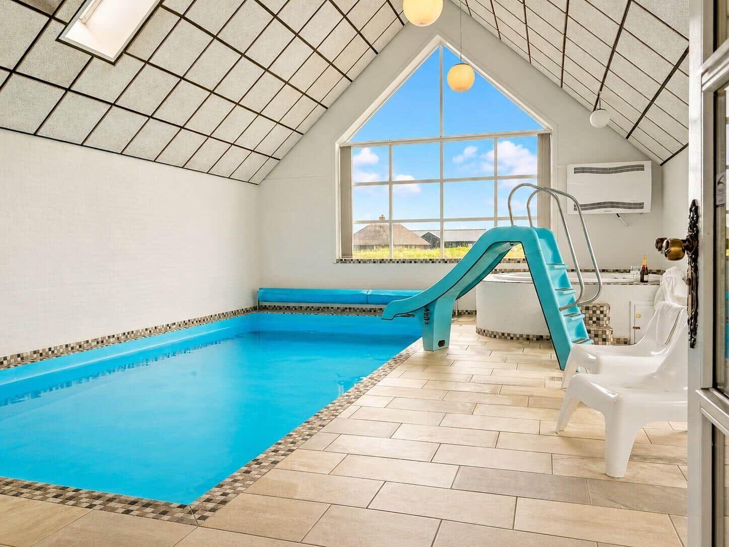 Innenpool mit blauer Rutsche und großem Fenster. Weiße Stühle und Fliesenboden.