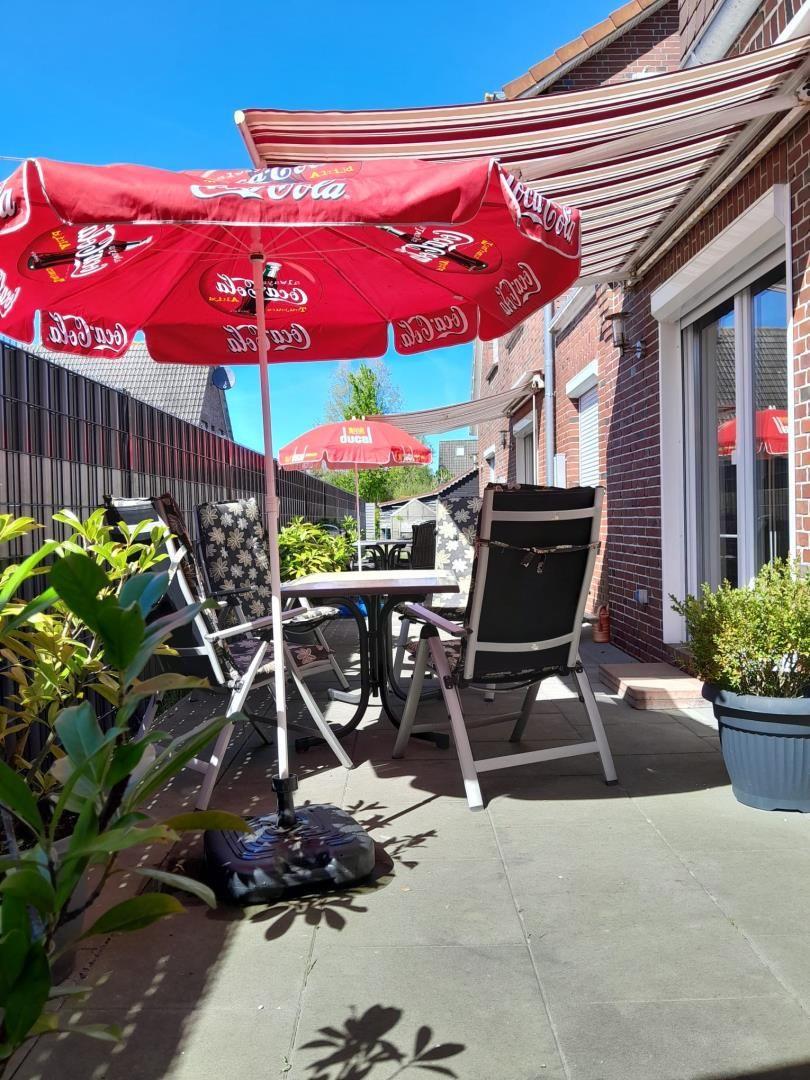 Terrasse mit Tisch, Stühlen und Coca-Cola-Sonnenschirm. Hintergrund: Haus und Pflanzen.