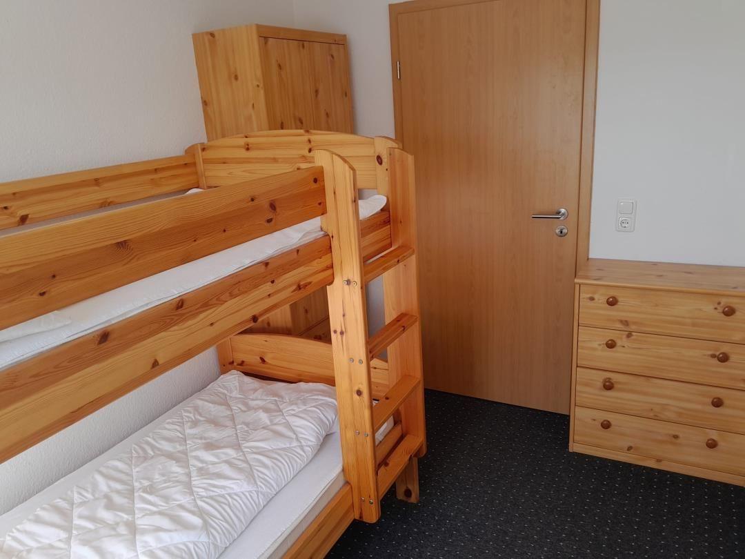 Zimmer mit Holz-Etagenbett, Schrank und Schrank. Tür und Steckdose sichtbar.
