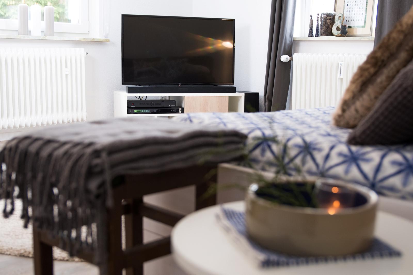 Ein Wohnzimmer mit Fernseher, Couch und Tisch. Auf dem Tisch steht eine Kerze.