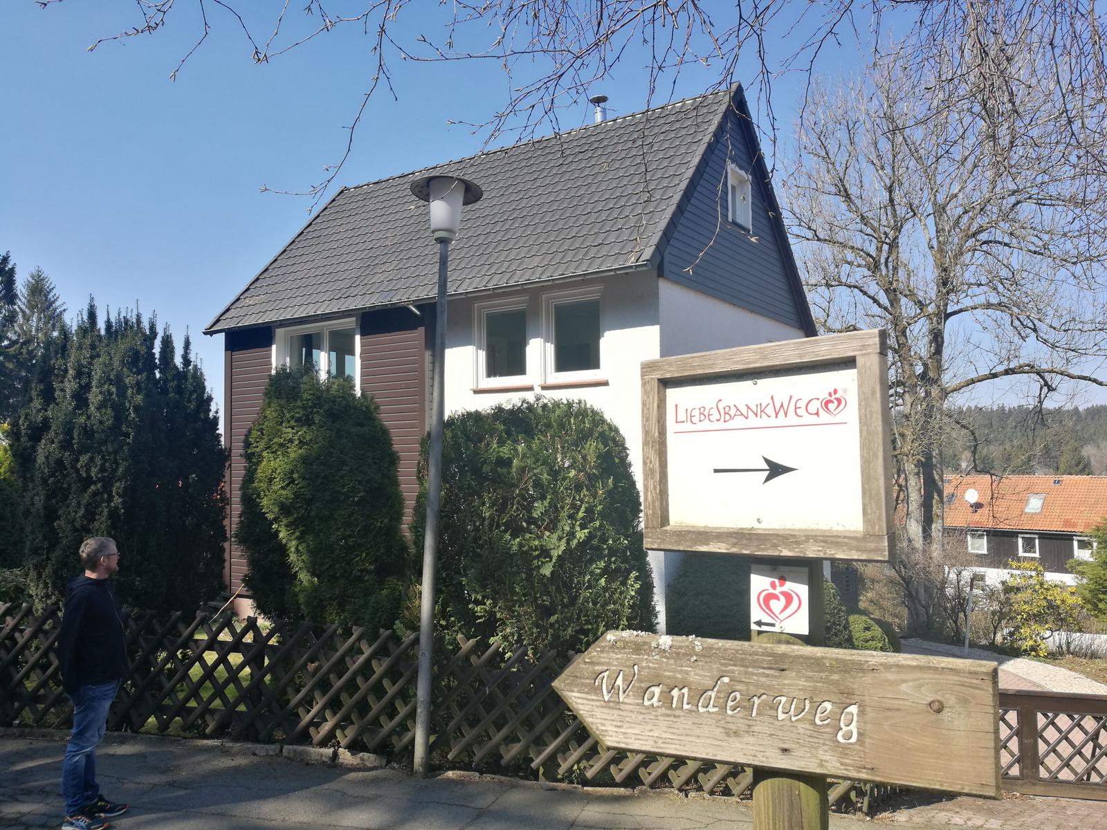 Haus mit Wanderweg-Schild und Liebesbankweg-Zeichen. Holzumzäunung und Bäume im Hintergrund.