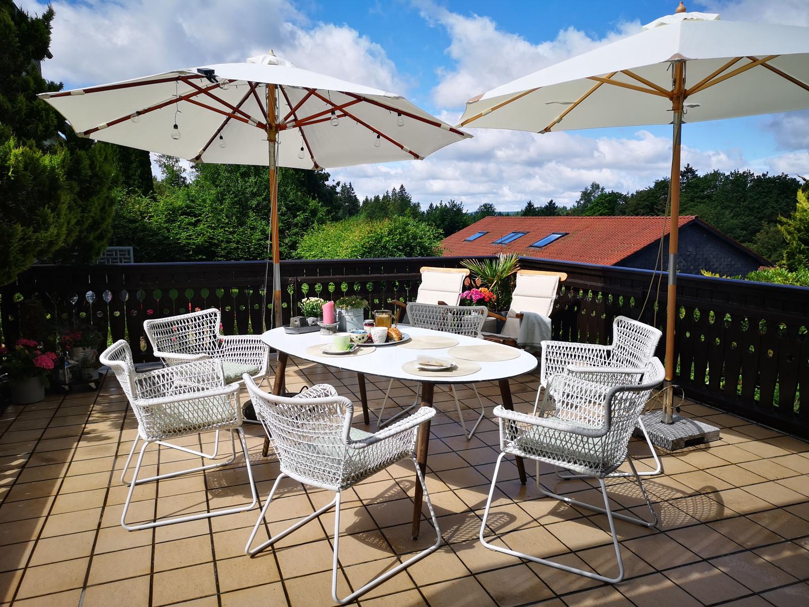 Terrasse mit weißem Tisch, Stühlen und Sonnenschirmen. Hintergrund: Wald und Haus mit rotem Dach.