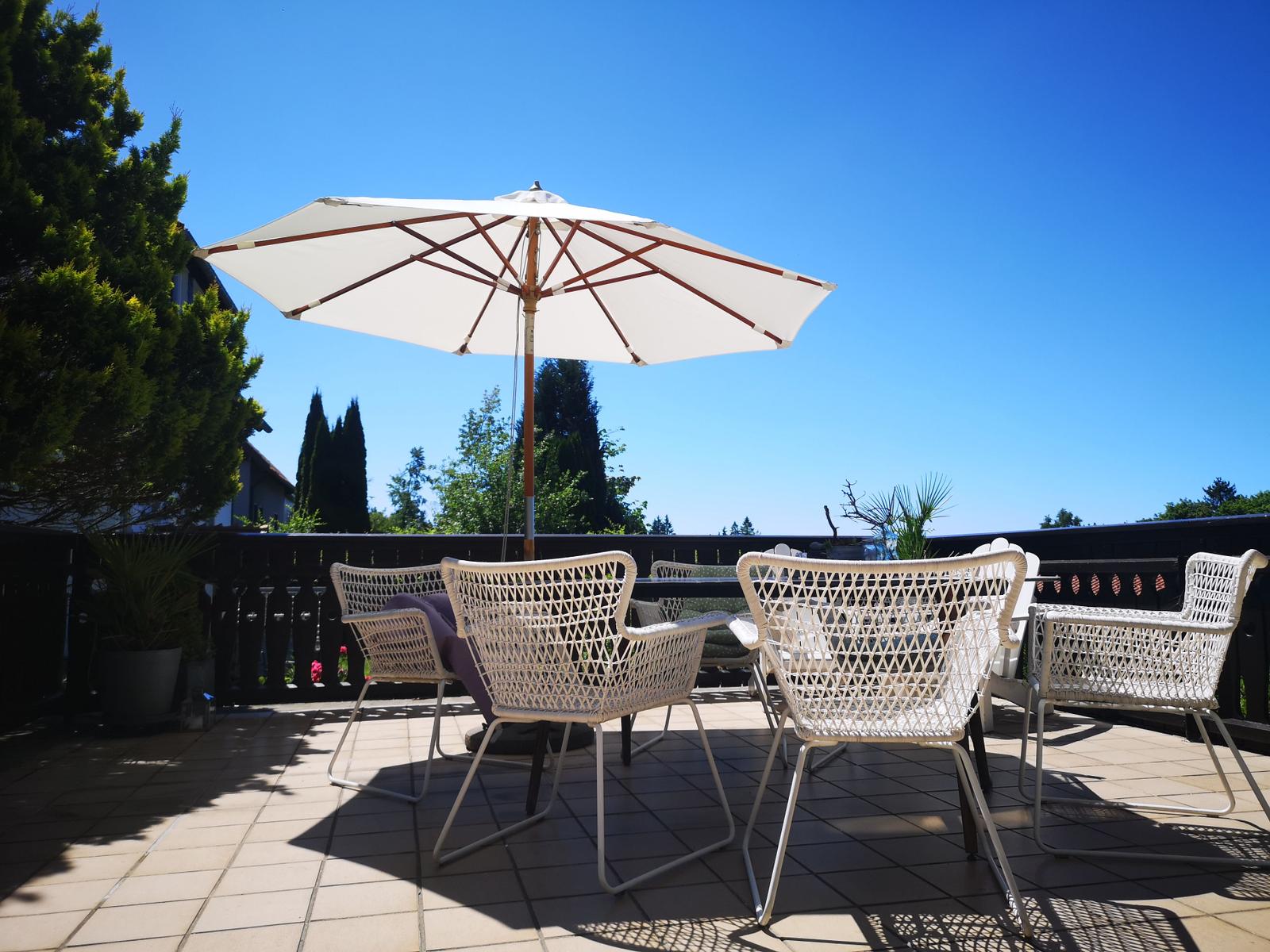 Terrasse mit weißem Sonnenschirm und Stühlen. Blick auf Bäume und klaren Himmel.