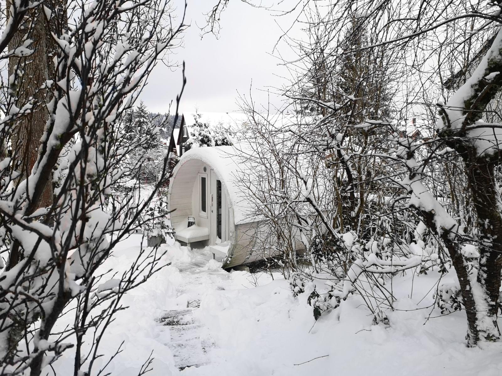 Weißer, zylindrischer Holzschuppen im Schnee, umgeben von schneebedeckten Bäumen.