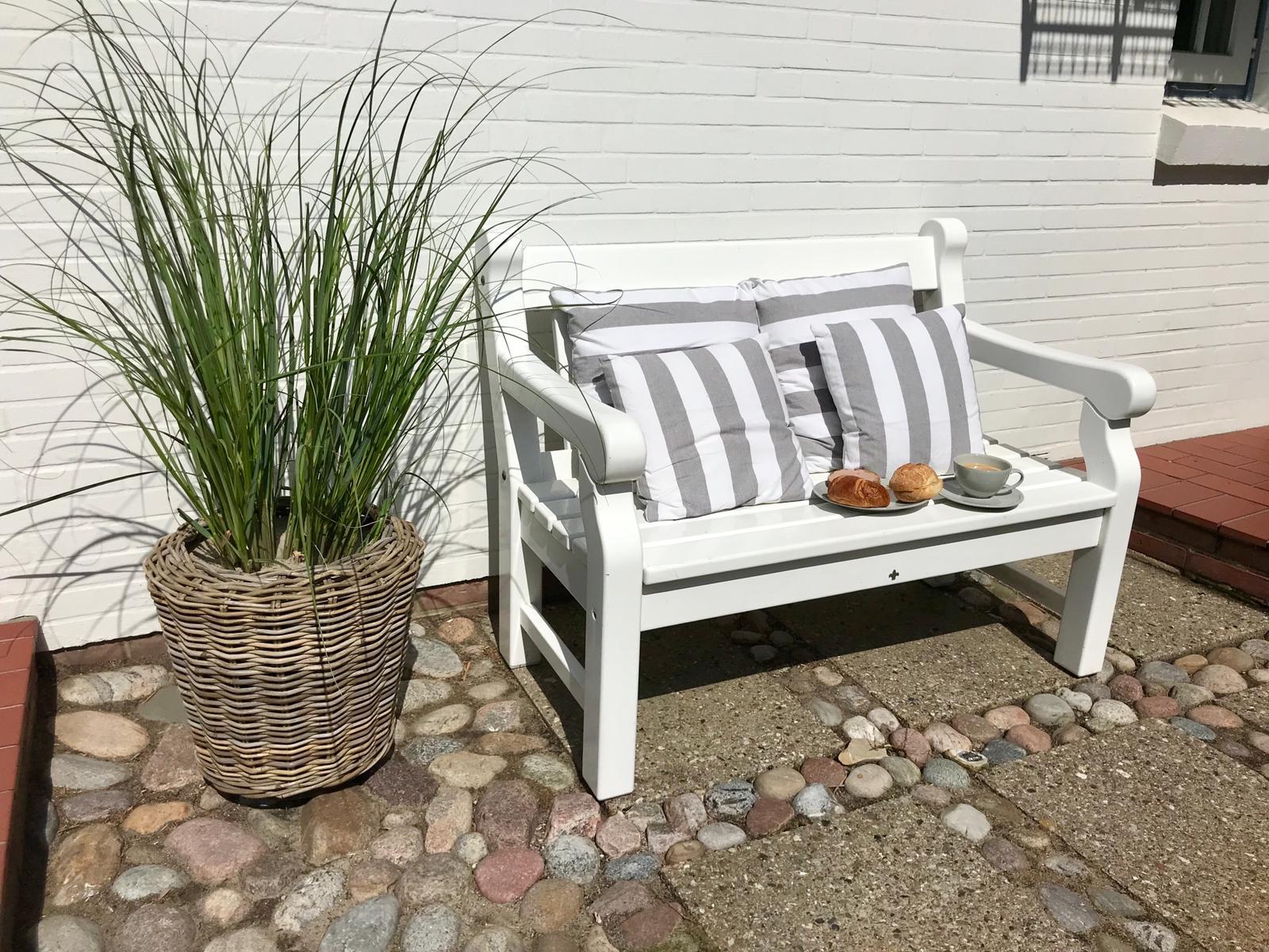 Weißer Gartenbank mit Streifenkissen, Kaffee und Brötchen, neben Pflanze in Korb.