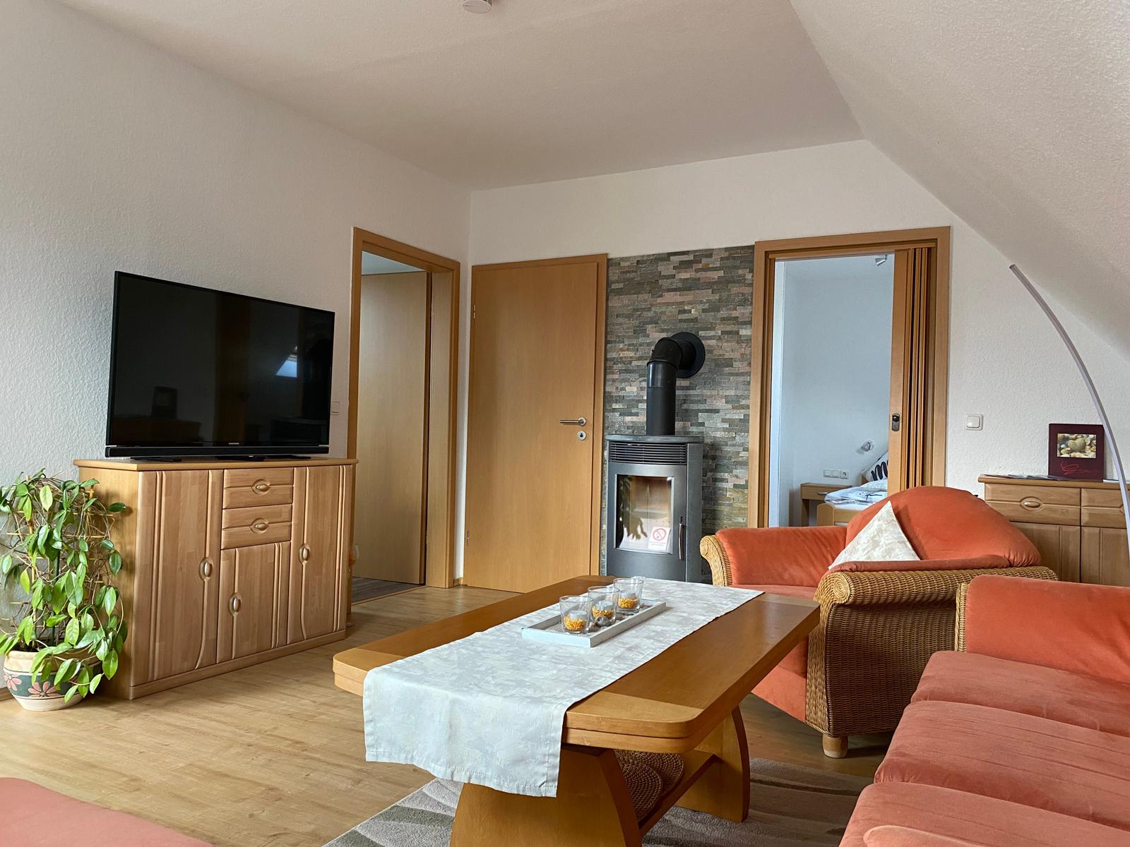Wohnzimmer mit Holzmöbeln, Fernseher und Kaminofen. Orange Sessel und Couch.